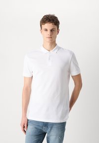 Camisa polo blanca de algodón con cuello y tapeta de tres botones, con un discreto logo bordado en el pecho. Jeans de color azul claro.
