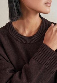 Pull en tricot marron foncé avec un col rond côtelé et des poignets, présentant une texture douce et une coupe décontractée, mettant en valeur des détails en tricot complexes.