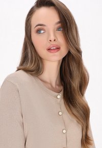 Bežový strukturovaný kardigan s kulatým výstřihem a čtyřmi dekorativními knoflíky v přední části. Dlouhé vlnité vlasy rámují obličej.