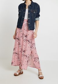 Jeansjacke mit hochgekrempelten Ärmeln über einem rosa floral gemusterten Maxikleid, das einen hohen Schlitz und weiße Sandalen mit einer massiven braunen Sohle hat.