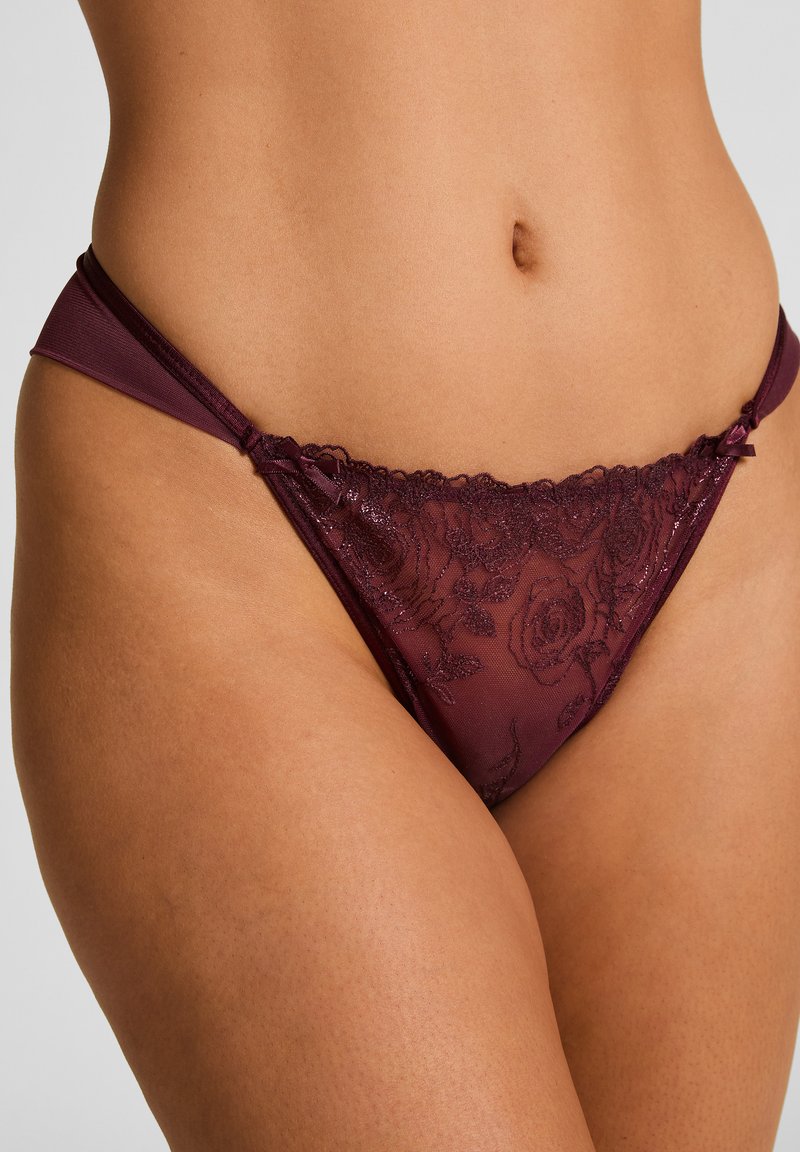 Burgunderfarbener Spitze-Tanga mit floralem Muster, bestehend aus einem durchsichtigen Material, zarter Borte entlang der Taille und dünnen Stoffträgern.