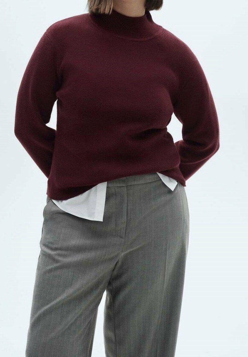 Pull côtelé bordeaux à col montant, porté par-dessus une chemise blanche à col, associé à un pantalon gris clair cintré.