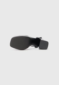 Sandal de pele preta com um salto quadrado e sola texturizada, vista pela parte de baixo contra um fundo branco liso.