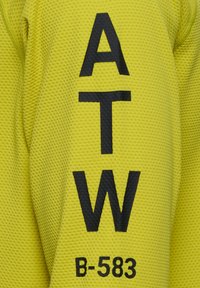 Helles gelbes Netzshirt mit fetter schwarzer Schrift "ATW" und "B-583", die auf der Vorderseite gedruckt ist. Strukturiert Oberfläche mit atmungsaktivem Design.