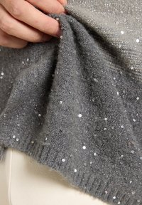Grijze trui met pailletten omhooggehouden, met een zachte textuur, fonkelende versieringen en geribbelde zoom. Duidelijke details van de stofsamenstelling zichtbaar.