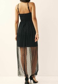 Schwarzes Kleid mit tailliertem Oberteil und verstellbaren Trägern, das mit einem透sichtigen, gepunkteten Überzug versehen ist, der bis zu den Knöcheln fällt. Offener Rückendesign.