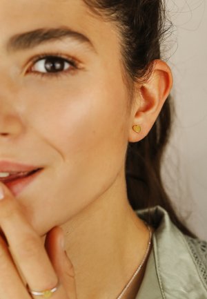 Close-up of woman with brown eyes wearing small gold heart-shaped earring, silver ring with green stone, and light green top.