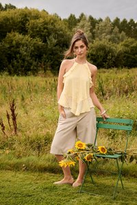 Jeune femme en haut jaune sans manches et culottes beiges se tient près d'une chaise verte tenant des tournesols dans un champ herbeux avec des arbres derrière.