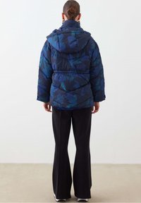 Giacca imbottita blu navy con grande stampa floreale in turchese e verde, dotata di cappuccio e polsini elasticizzati, indossata sopra pantaloni neri.