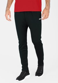 Schwarze Sporthose mit schmalem Schnitt, aus weichem Material. Merkmale sind das weiße "JAKO"-Logo, der elastische Bund und passende schwarze Sneaker.