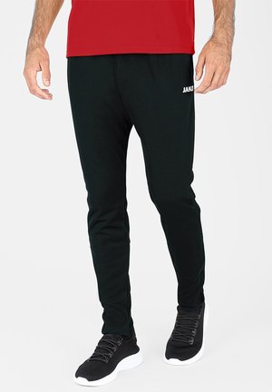Pantalons de sport noirs à coupe fuselée, fabriqués en matériau doux. Dotés d'un logo "JAKO" blanc, d'une taille élastique et accompagnés de chaussures de sport noires assorties.