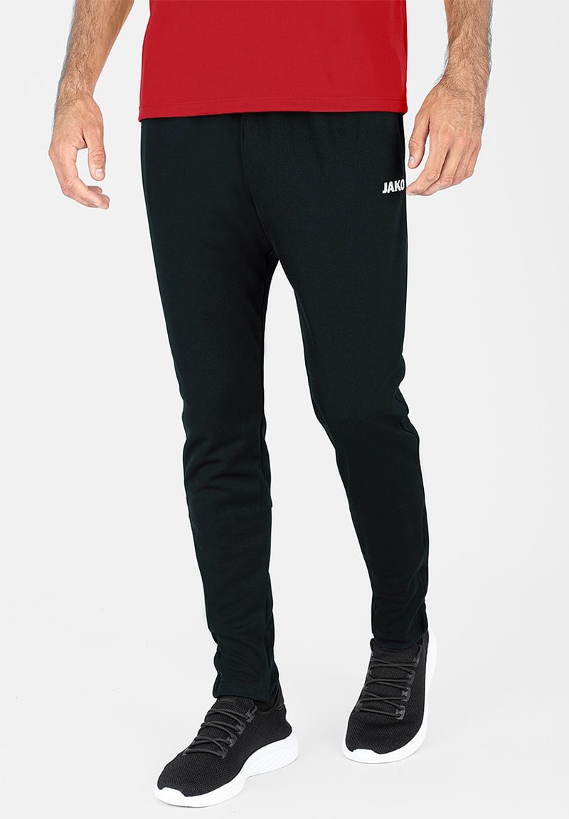 Schwarze Sporthose mit schmalem Schnitt, aus weichem Material. Merkmale sind das weiße "JAKO"-Logo, der elastische Bund und passende schwarze Sneaker.