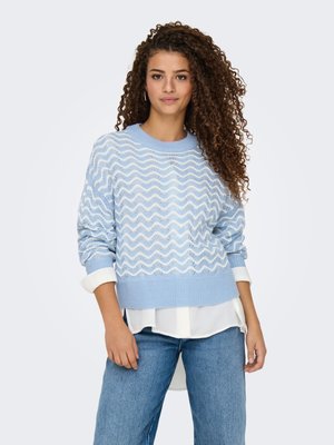 Donna con capelli ricci indossa un maglione azzurro chiaro con motivo sopra una camicia bianca e jeans blu, in piedi contro uno sfondo chiaro uniforme.