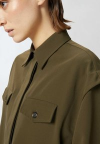 Giacca camicia verde oliva con colletto, due tasche sul petto con bottoni e una texture liscia. Vestibilità sagomata, dettagli di design minimalisti.