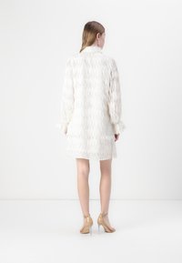 Manteau blanc texturé avec un design à franges, manches longues et une silhouette légèrement cintrée, associé à des talons hauts nude.