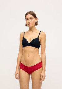 Sutien wireless negru cu decolteu adânc și bretele subțiri, asortat cu chiloți roșii cu dantelă, având un detaliu mic sub formă de fundiță.