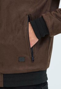 Braune Wildlederjacke mit einer Reißverschlusstasche, gerippten schwarzen Bündchen und einem kleinen schwarzen Logo-Patch nahe der Taille. Weicher Stoff, elegantes Design.