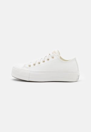 Hvid lavtop canvas sneaker med gummisål og metaløjer, vist fra siden mod en ensfarvet baggrund.