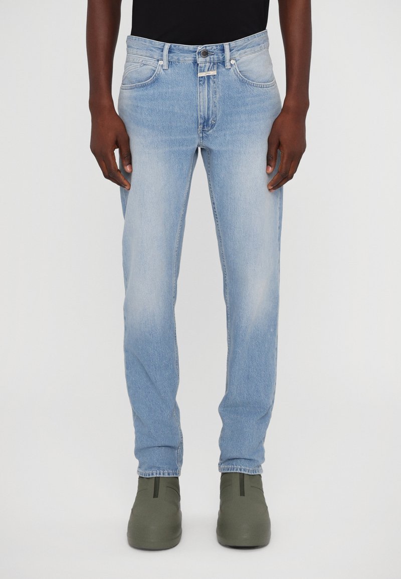 Jean en denim bleu clair avec une coupe droite, cinq poches, un dégradé subtil et une fermeture à bouton en métal. Associé à des chaussures vertes à enfiler.