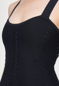 Fekete, testhezálló felső vékony pántokkal, texturált anyagból, és függőleges kivágási részletekkel az oldalakon, amely közel áll a testhez.