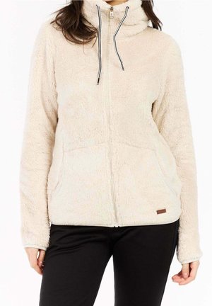 Kobieta w kremowej, puszystej kurtce na zamek z przodu z kieszeniami i czarnymi troczkami, połączonej z czarnymi spodniami.