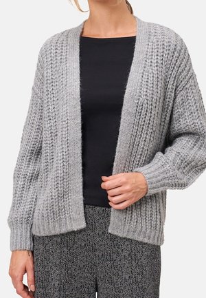Femme portant un cardigan épais gris clair en maille sur un haut noir et un pantalon gris à motifs, tenant le bord du cardigan d'une main.