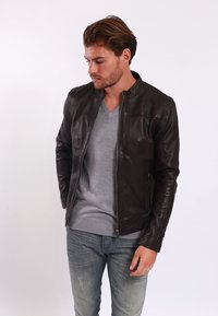 Giacca di pelle marrone con una vestibilità slim, colletto alzato e chiusura a zip. Il modello indossa un maglione grigio a scollo a V e jeans strappati.