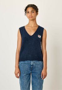 Marineblauwe, mouwloze trui met een V-hals ontwerp, voorzien van een zachte, fuzzy textuur en een kleine kleurpatch aan de linkerzijde.