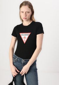 Cămășă neagră ajustată cu un logo triunghiular roșu și text "GUESS?", asociată cu blugi denim albastre. Design simplu, mâneci scurte și guler rotund.