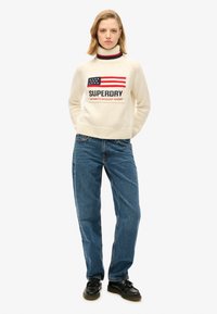 Maglione lavorato a maglia bianco con un design della bandiera americana in rosso e blu con il testo "SUPERDRY". Abbinato a jeans in denim blu e scarpe nere.