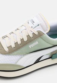 Gros plan d'une basket Puma avec des panneaux blancs, beiges et verts, des lacets crème, un tissu en mesh et le logo "Puma" sur le côté.