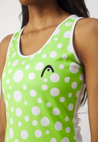Gros plan d'une femme portant un haut sans manches vert citron avec des pois blancs et un petit logo noir incurvé sur la poitrine.