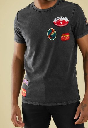 Mężczyzna noszący wyblakły czarny t-shirt z kolorowymi haftowanymi naszywkami, w tym "Ski Patrol", "Guess" i "Est. 1981", w połączeniu z czarnymi spodniami.