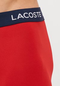 Calções atléticos vermelhos com uma textura suave, apresentando uma cintura azul-marinho com "LACOSTE" em letras brancas.