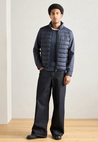 Marineblå puffer-vest over en ribstrikket marineblå sweater, kombineret med vidbenede mørk denimjeans og sorte sko, med et moderne, lagdelt look.