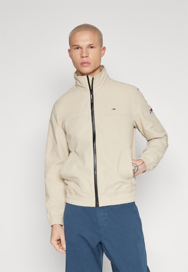 Bežna jakna na zip s crnim zatvaračem, visokim ovratnikom, elastičnim manžetama i pojasom. Logotip na lijevoj strani prsiju. Nosi se preko plavih hlača.