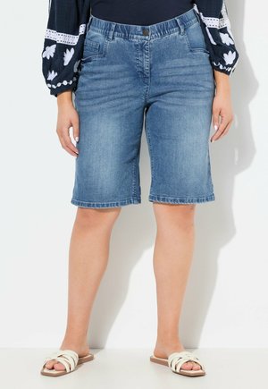 Bermuda shorts in denim blu medio, con vita media, design a cinque tasche e orlo sfrangiato. Indossati con sandali di colore chiaro.