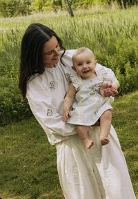 Bijela vezeno bluzica s dugim rukavima, uparena s svijetlim slobodnim hlačama. Beba u prugastom kombinezonu s volanima, smiješi se. Zelena pozadina.