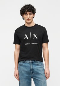 Svart bomulls-t-skjorte med korte ermer, med en hvit grafisk logo "A/X" og teksten "ARMANI EXCHANGE" i midten. Brukt sammen med blå jeans.