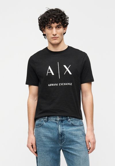 T-shirt noir en coton à manches courtes, avec un logo graphique blanc "A/X" et le texte "ARMANI EXCHANGE" au centre. Porté avec un jean bleu.