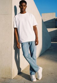 Jeune homme en t-shirt blanc, jean bleu clair et baskets blanches, appuyé contre un mur en béton sous un ciel bleu clair.