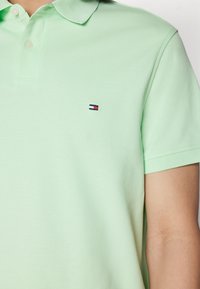 Světle zelené polokošile s malým obdélníkovým výšivkovým logem v červené, bílé a námořnické modři na hrudi, kterou nosí osoba.