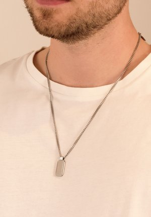 Silberne Halskette aus Edelstahl mit einem rechteckigen Anhänger, an einer dünnen Kette, die über einem schlichten weißen T-Shirt getragen wird.