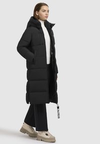 Giacca a vento nera con cappuccio, caratterizzata da un design trapuntato, zip anteriore e dettagli con cordoncino. Indossata sopra una maglietta bianca e pantaloni neri.