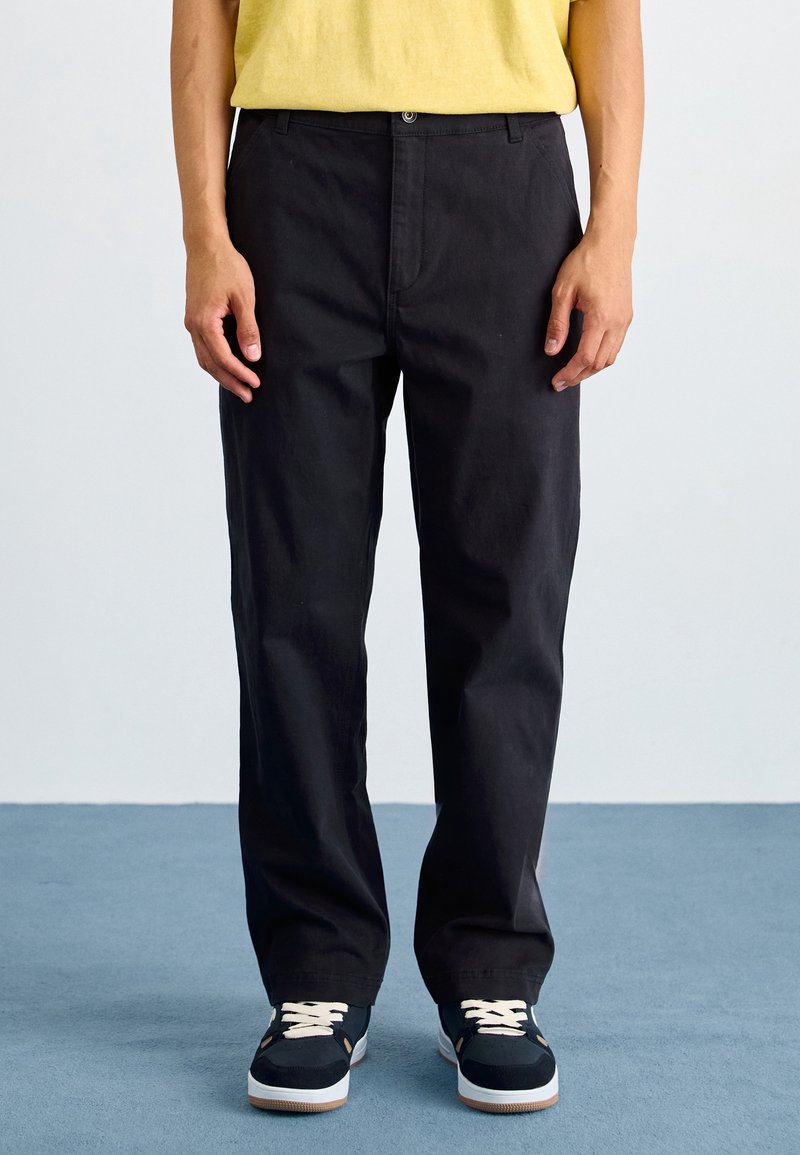 Svarta bomullsbyxor med rak passform, med framfickor och knappstängning. Matchas med marinblå sneakers och en gul skjorta.