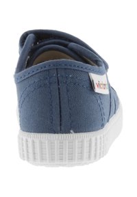 Sneaker in tela blu con suola in gomma bianca e cinturino in velcro, vista da dietro, con una piccola etichetta "victoria" multicolore.