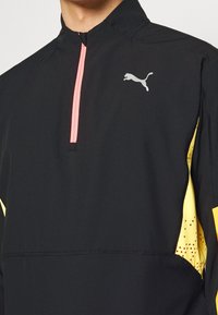 Giacca nera con zip a quarto di lunghezza, zip rosa, pannelli laterali gialli e logo grigio. Texture liscia con leggere perforazioni vicino all'orlo.