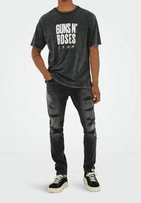 Jeans noirs déchirés avec des trous et un t-shirt noir à motif graphique affichant "GUNS N' ROSES" en grandes lettres blanches avec des accents d'étoiles. Baskets noir et blanc.