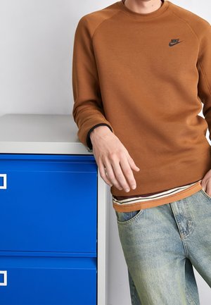 Person trägt braunen Nike-Sweatshirt und hellblaue Jeans, lehnt an einem weißen und blauen Aktenschrank.