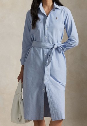 Vrouw draagt een lichtblauwe blousejurk met knopen en een taillestrop, houdt een witte handtas vast, staand tegen een neutrale achtergrond.